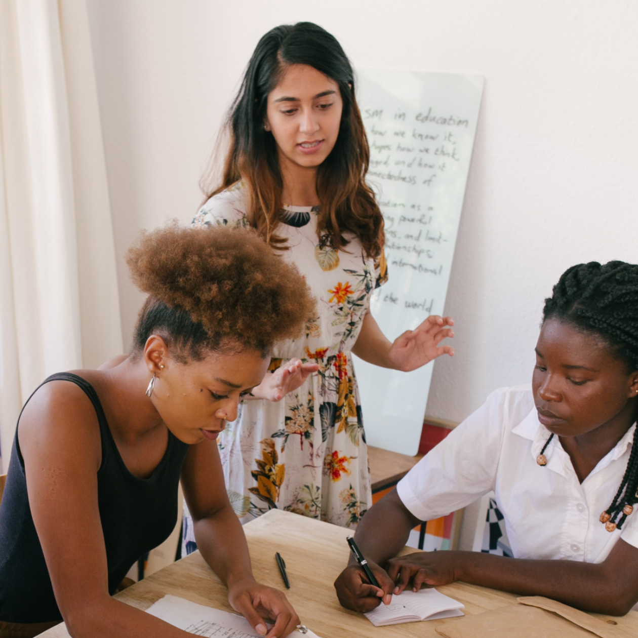 A photo of two people sitting at a desk writing on paper, and one person standing behind them taking to them.